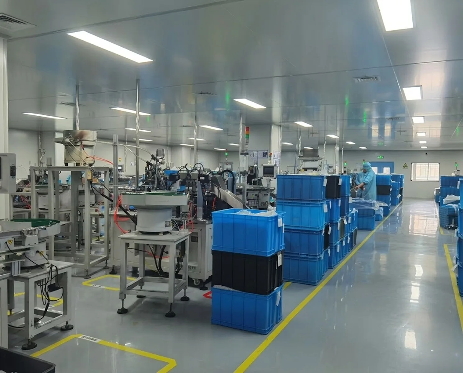 Workers in protective suits operating automated assembly machines for skincare packaging inside a dust-free cleanroom with stacked blue crates