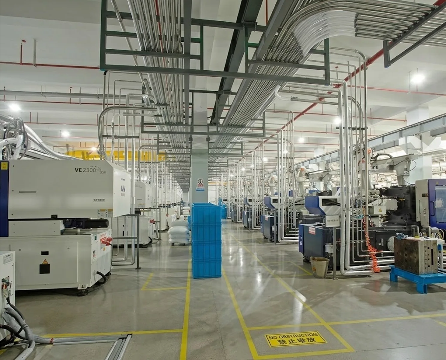 Wide view of a cosmetic packaging factory floor showing complex overhead metal piping and rows of heavy injection molding machines