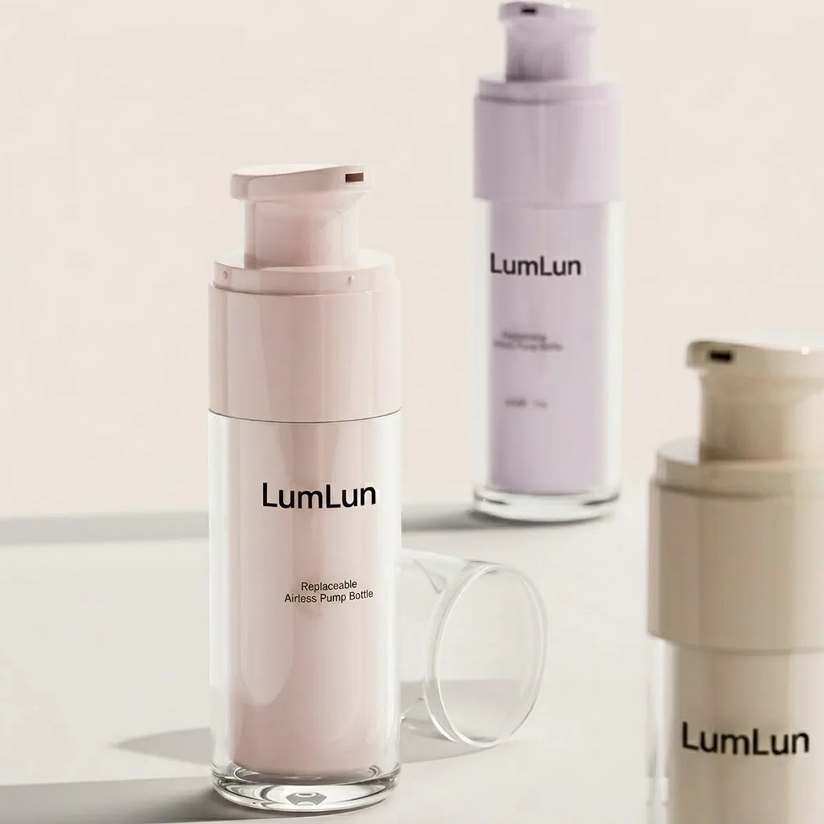 Three LumLun clear PS outer shell replaceable airless bottles in lifestyle display — blush pink in foreground with LumLun branding and Replaceable Airless Pump Bottle label, lavender in background, and warm beige partially visible at right, clear PS outer shell showing colored PP inner core, OEM custom serum lotion packaging