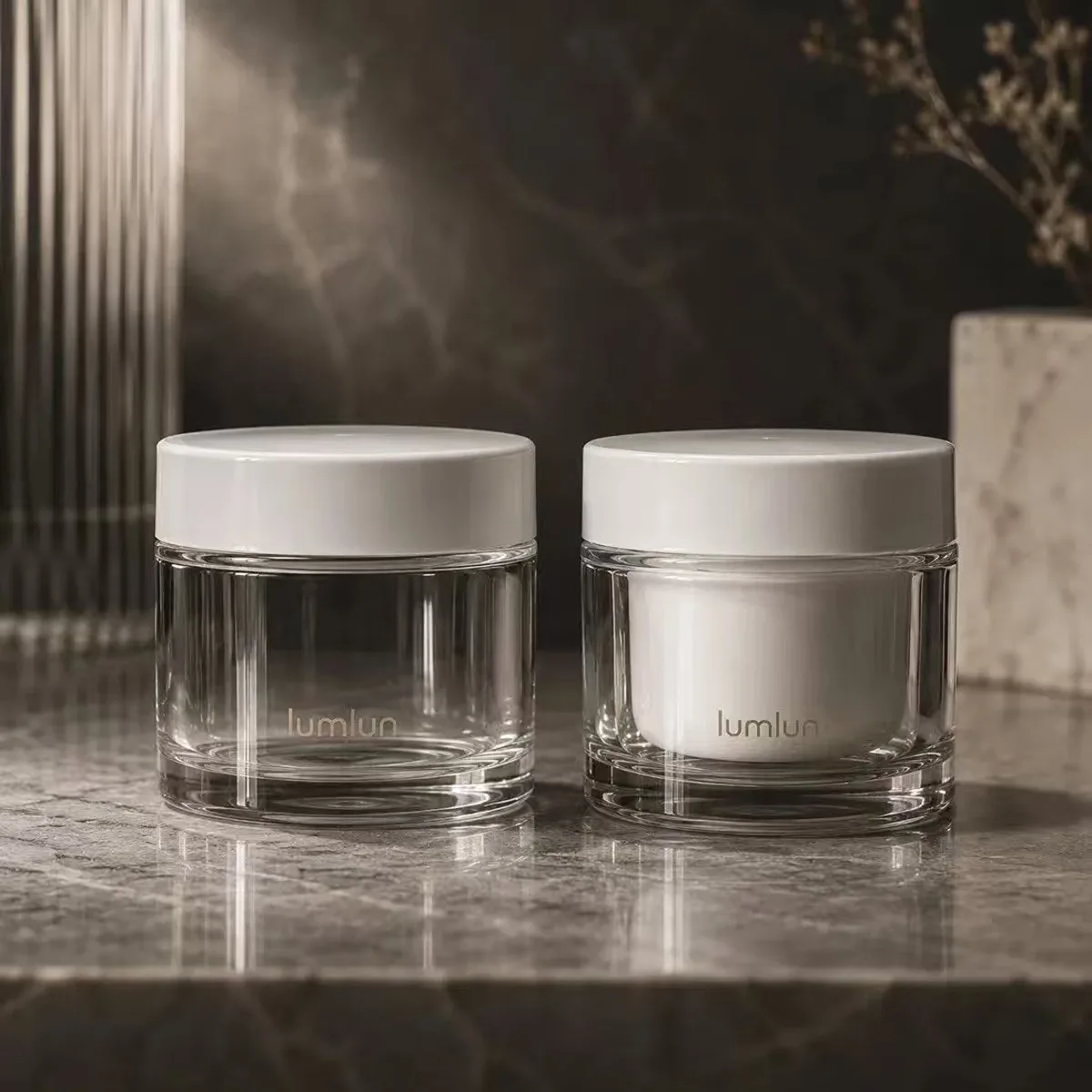 Two clear PETG cream jars on dark marble — one empty showing inner bottle structure, one filled with white cream