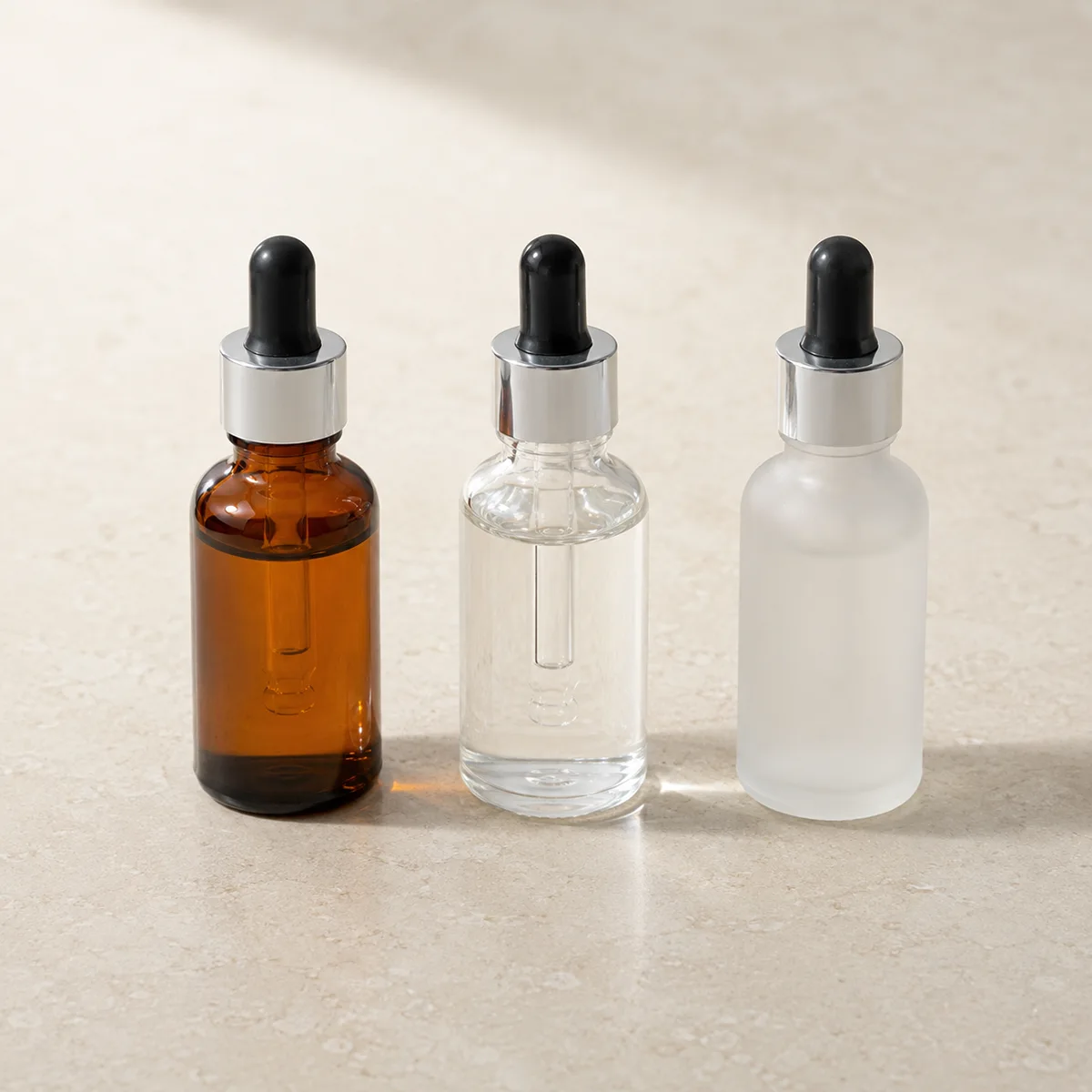 Three skincare dropper bottles in amber glass clear glass and frosted finish displayed on white surface showing packaging options for skincare brands