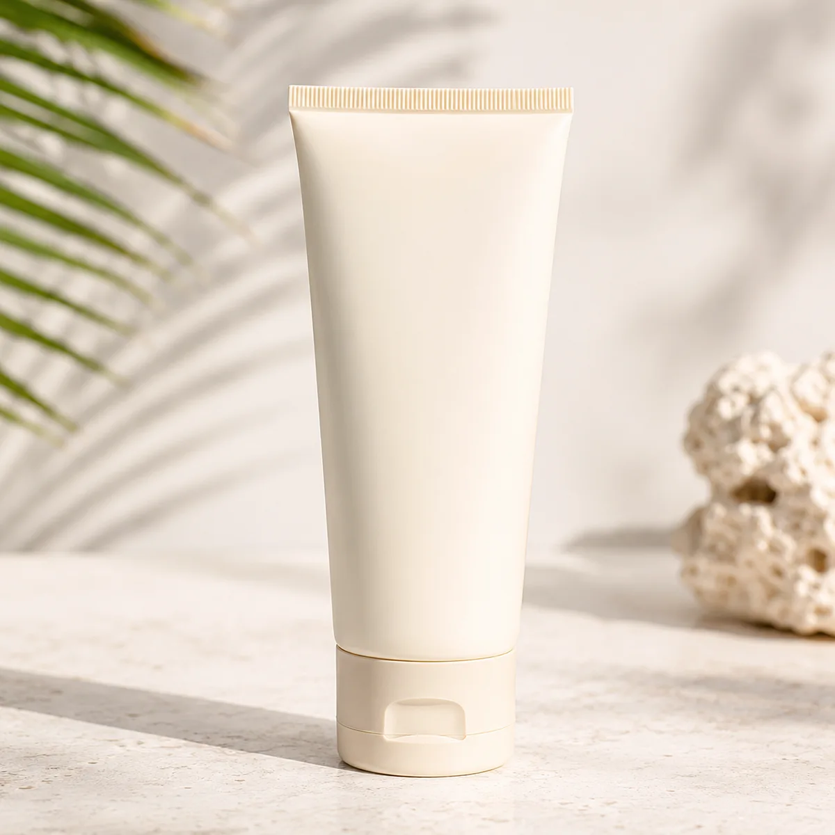 Cream-colored sunscreen squeeze tube with a flip-top cap on a sandy beach-style background with palm shadow