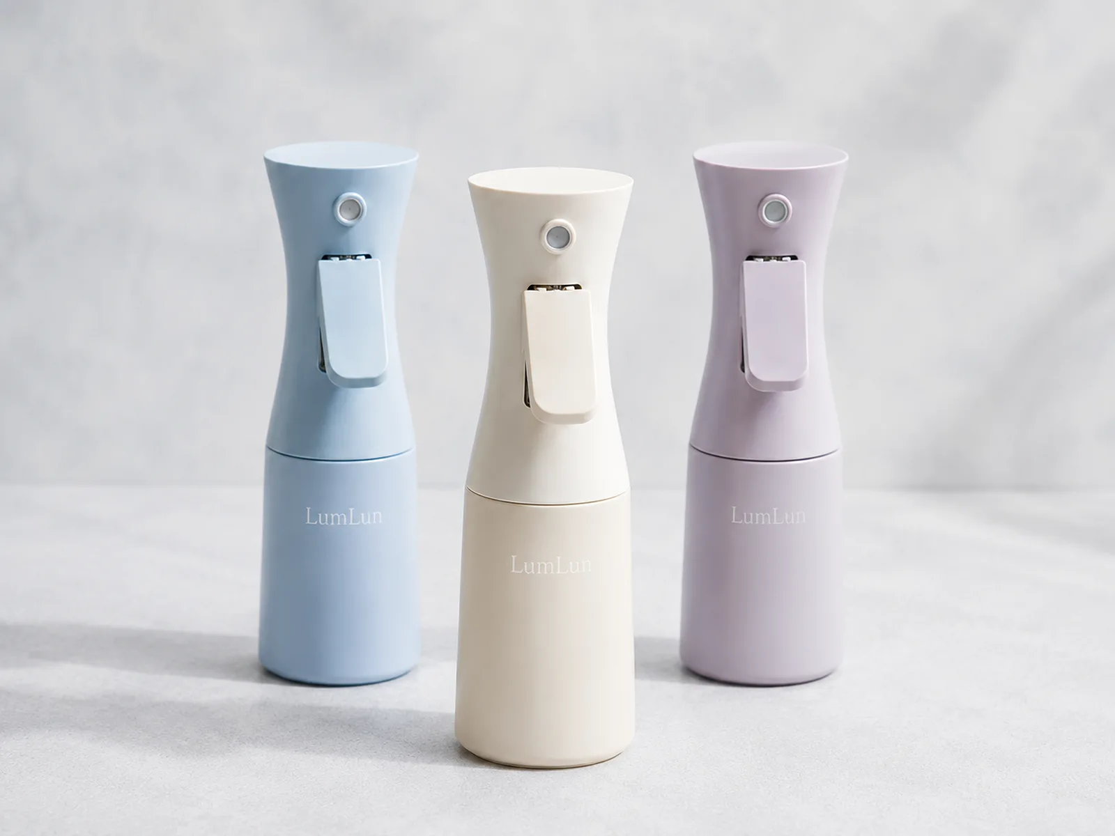 Three LumLun continuous mist spray bottles in pastel blue, ivory and lavender with ergonomic trigger designs