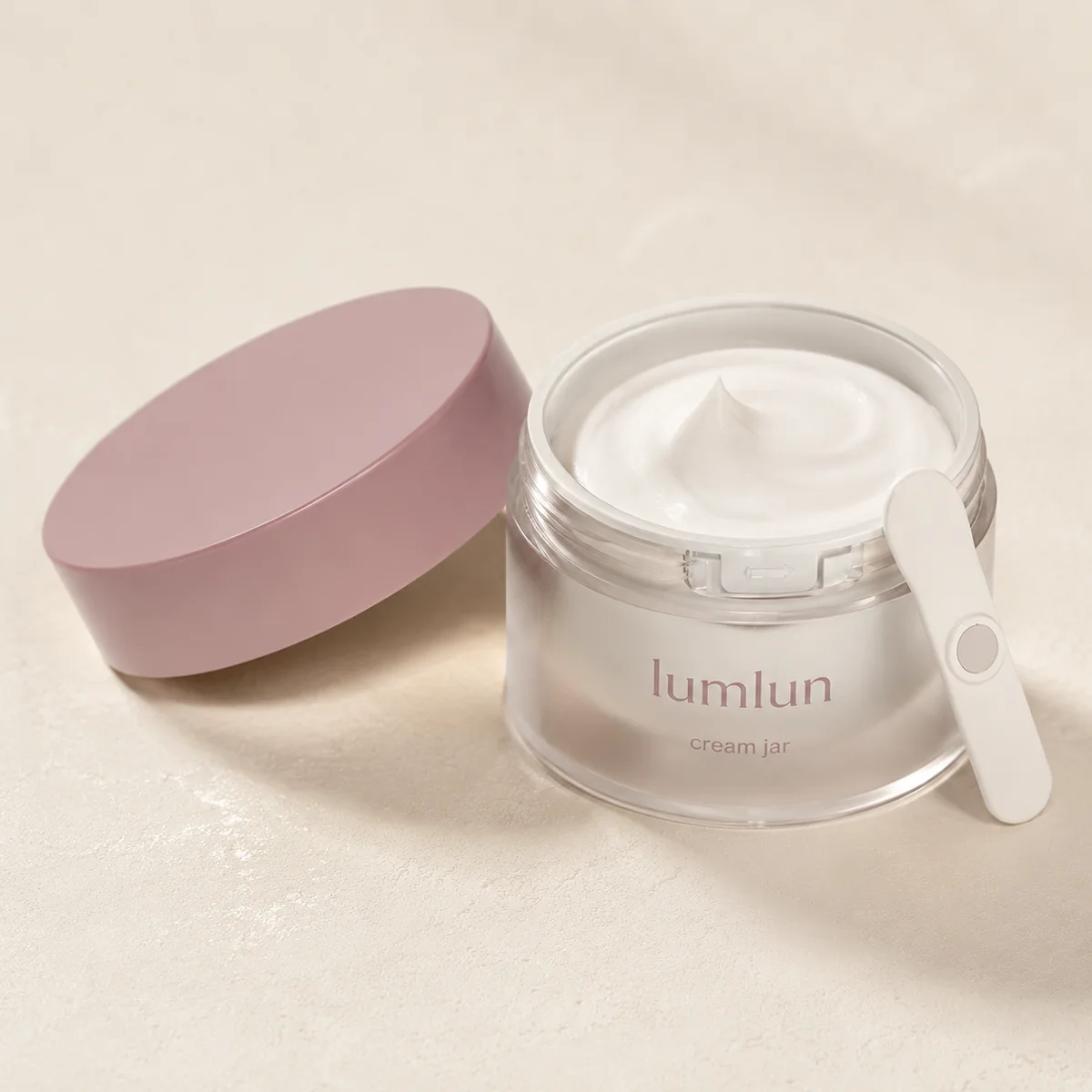 Open Lumlun cream jar with a pink lid, white cream, and magnetic spatula on beige surface.