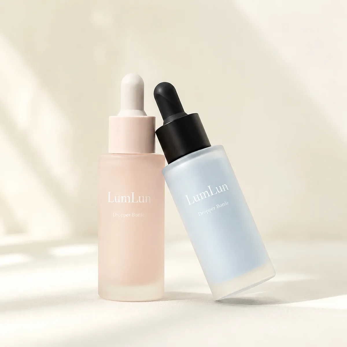 Two Lumlun frosted glass dropper bottles in peach and blue, one upright and one leaning beside it.